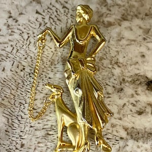May include: A gold-toned metal brooch depicting a figure in a long dress with a bow at the waist, standing next to a dog. A thin gold chain connects the figure's hand to the dog's collar, creating a linked design in a polished finish.