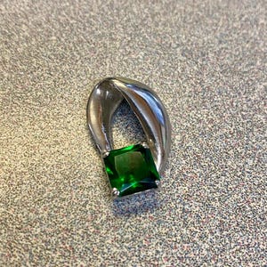 May include: A silver-toned metal pendant featuring a single, square-cut, deep green gemstone held in a four-prong setting. The metal frame curves upward in a smooth, polished, teardrop-like shape that wraps around the top of the stone.