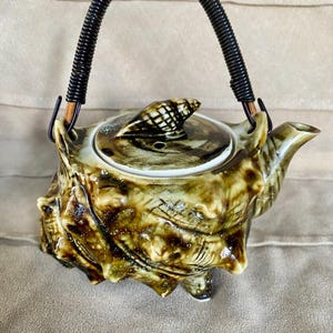 May include: A ceramic teapot shaped like a textured seashell with a glossy brown and green glaze. The lid features a small shell finial, and the vessel includes a curved spout and a black coiled wire handle attached to two upright lugs.