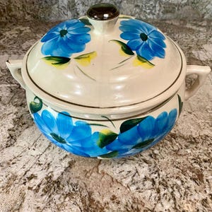 May include: A round ceramic tureen with a lid, featuring a cream-colored base decorated with hand-painted bright blue flowers, green leaves, and yellow accents. The tureen has two side handles and a dark brown knob on the center of the lid.