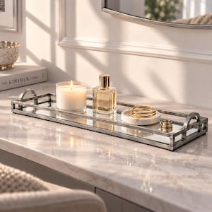 May include: A rectangular mirrored vanity tray with a silver-toned metal frame and arched handles. The tray holds a white candle in a glass jar, a clear glass perfume bottle with a gold cap, a small white marble dish, gold bangles, and a gold ring.