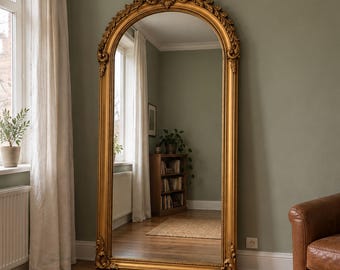 May include: A tall, floor-standing mirror with an ornate, arched gold frame featuring intricate carved detailing at the top and bottom. The mirror has a reflective glass surface that shows a room interior, including a wooden bookshelf and flooring.