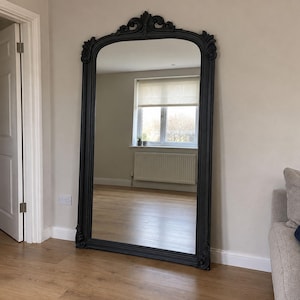 May include: A large, floor-standing mirror featuring a matte black frame with an arched top and ornate, carved detailing at the crown and bottom corners. The rectangular glass surface reflects a room with a window, a radiator, and light flooring.