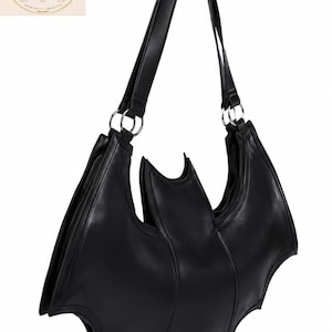 Puede incluir: Un bolso de hombro negro con forma de murciélago y acabado liso. El bolso presenta una sección central con extensiones puntiagudas en forma de ala a ambos lados y una correa para el hombro sujeta con anillas de metal de tono plateado.
