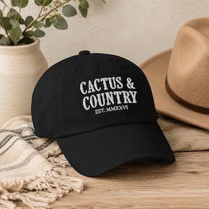 Może przedstawiać: Czarna czapka z daszkiem z białym haftowanym napisem CACTUS & COUNTRY EST. MMXXVI. Czapka posiada usztywnianą koronę, zakrzywiony daszek oraz guzik na górze. Haft znajduje się na środku przedniego panelu czapki.