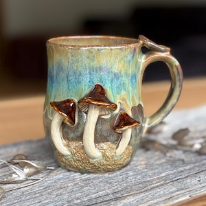 Può includere: Una tazza in ceramica con smalto lucido nei toni del blu, verde e marrone. L'esterno presenta tre funghi scolpiti in rilievo con cappelli marrone scuro e gambi color crema, su una base strutturata che ricorda il muschio del sottobosco.