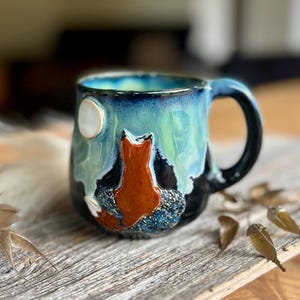 Puede incluir: Una taza de cerámica con un esmalte brillante en tonos azules y verdes. El exterior muestra un zorro naranja esculpido en relieve, sentado y mirando hacia una luna circular blanca sobre un fondo oscuro y texturizado, con un asa curva.