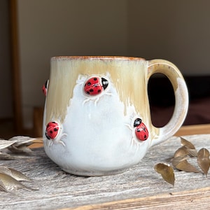 Peut inclure: Une tasse en céramique avec un vernis bicolore, présentant une partie supérieure beige chaud et une base blanche. L'extérieur est orné de coccinelles rouges en relief peintes à la main avec des points noirs, réparties sur le corps de la tasse.