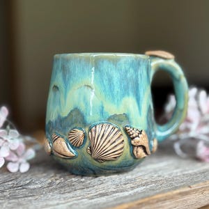 Op de afbeelding: Een keramische mok met een glanzend blauwgroen glazuur en verhoogde, getextureerde schelpversieringen rond de basis. De mok heeft een stevig handvat en een glad, afgerond lichaam, met een mix van blauwe en groene tinten over het geheel.