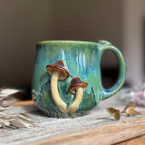 Könnte beinhalten: Ein Keramikbecher mit einer glänzenden grün-blauen Glasur, der auf der Vorderseite zwei erhabene, dreidimensionale Pilzskulpturen aufweist. Die Pilze haben helle Stiele und braune Kappen. Der Becher hat einen geschwungenen Henkel an der rechten Seite.
