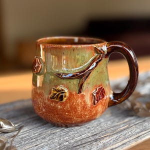 Może przedstawiać: Ceramiczny kubek z błyszczącym szkliwem w odcieniach szałwiowej zieleni i palonej pomarańczy. Zewnętrzna strona ozdobiona jest trójwymiarowymi detalami w formie ciemnobrązowej gałęzi drzewa oraz kilkoma małymi liśćmi rozrzuconymi na powierzchni.