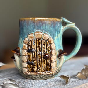 Peut inclure: Une tasse en céramique avec une finition émaillée bleue et verte, ornée d'une porte en bois marron sculptée en relief avec une arche en pierre. La tasse présente de petits champignons marron sur les côtés et une anse incurvée, offrant un aspect texturé en trois dimensions.