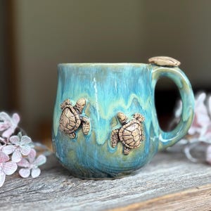 Könnte beinhalten: Ein Keramikbecher mit einer glänzenden, blau-grünen Tropfglasur. Der Becher ist mit zwei modellierten, hellbraunen Meeresschildkröten auf der Außenseite verziert. Auf dem Henkel befindet sich ein kleiner, flacher, hellbrauner Schildkrötenpanzer.