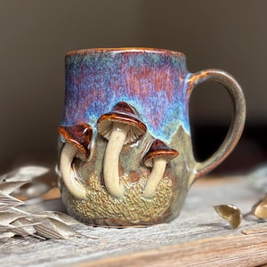 Könnte beinhalten: Ein Keramikbecher mit einem handgeformten Relief von drei Pilzen auf der Vorderseite. Der Becher hat eine glänzende Glasur mit einem Farbverlauf von tiefem Blau und Violett oben, der in eine strukturierte, erdfarbene grün-braune Basis übergeht.