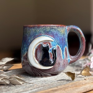 May include: A ceramic mug with a glossy glaze featuring a blend of blue, purple, and brown tones. The mug has a raised, three-dimensional relief of a black cat sitting inside a white crescent moon shape on the front surface and a curved handle.