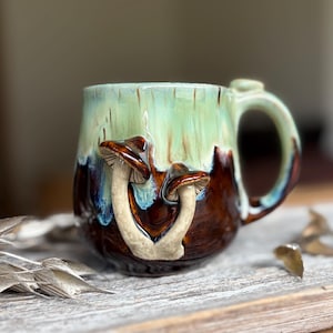Può includere: Una tazza in ceramica con smalto bicolore, caratterizzata da una parte superiore verde menta chiaro e una base marrone cioccolato intenso. Due funghi scolpiti in rilievo con gambi beige e cappelli marroni sono applicati sulla parte frontale.