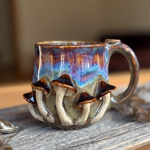 Pode incluir: Uma caneca de cerâmica com um esmalte brilhante em tons de azul profundo, roxo e castanho. O exterior é decorado com vários cogumelos esculpidos em três dimensões com chapéus castanhos escuros e caules claros à volta da base.