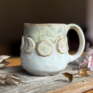 May include: A ceramic mug with a rounded body and a single handle, featuring a two-tone glaze of pale green at the top and off-white at the bottom. The exterior is decorated with a series of raised, embossed moon phase symbols encircling the center.