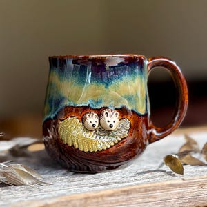 Puede incluir: Una taza de cerámica con un esmalte brillante que presenta un degradado de marrón oscuro, azul verdoso y amarillo. La taza tiene un relieve esculpido de dos pequeños erizos descansando sobre una hoja de helecho texturizada en la parte frontal.