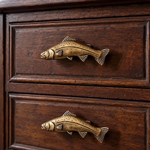 May include: Two brass-toned metal drawer pulls shaped like fish, each featuring detailed scales, fins, and a spotted pattern. These horizontal handles are mounted onto the front of two stacked dark brown wooden drawers with a rectangular frame design.