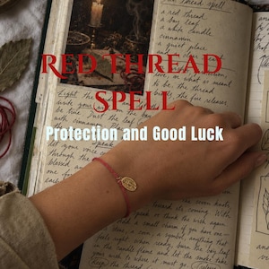 May include: A thin red string bracelet featuring a small, oval-shaped gold-toned metal charm. The bracelet is displayed against an open book with handwritten text titled RED THREAD SPELL and the subtitle Protection and Good Luck.