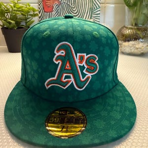 Op de afbeelding: Een groene honkbalpet met een herhalend klaverbladpatroon en het A's-logo van de Oakland Athletics geborduurd in wit, groen en oranje op de voorkant. Een ronde goudkleurige en zwarte sticker op de platte klep toont de maat 7 3/8.
