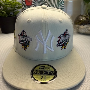 Op de afbeelding: Een lichtbeige New Era 59FIFTY pet met een wit geborduurd New York Yankees logo op de voorzijde, geflankeerd door twee World Series patches. Een ronde zwart-gouden sticker op de platte klep geeft de maat 57,7 cm aan.