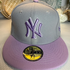 Könnte beinhalten: Eine graue Baseballkappe mit einem aufgestickten violetten New York Yankees Logo auf der Vorderseite. Die Kappe hat einen violetten Schirm, einen violetten Knopf oben und einen runden 59FIFTY-Aufkleber auf dem Schirm mit der Größe 57,7 cm.