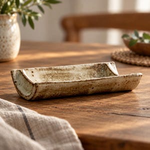 May include: A rectangular ceramic tray with a curved, trough-like shape. The piece features a speckled, off-white glaze with brown, earthy accents and a textured, uneven surface finish along the edges and interior base of the vessel.