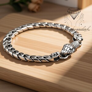 May include: A silver-toned metal chain bracelet featuring a snake head clasp and a small skull detail. The bracelet is composed of interlocking links with a polished finish, creating a continuous circular shape. The snake head is textured.
