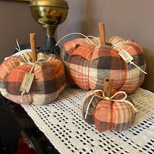 May include: Three fabric pumpkins in varying sizes featuring an orange, brown, and cream plaid pattern. Each pumpkin is topped with a brown cinnamon stick stem and tied with thin twine. Two of the pumpkins feature a small wooden tag labelled Linhas.