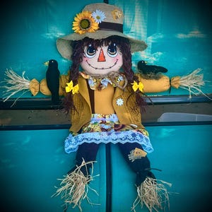 May include: A fabric scarecrow doll wearing a tan burlap hat decorated with a sunflower and daisy, a mustard yellow cardigan, and a skirt with a sunflower print and white lace trim. The doll has dark brown yarn hair, black birds on its shoulders, and straw accents at the wrists and ankles.