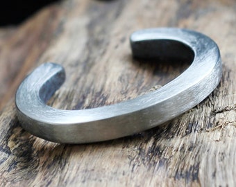Puede incluir: Un brazalete de metal plateado con acabado cepillado. El brazalete está abierto y tiene una forma redondeada.