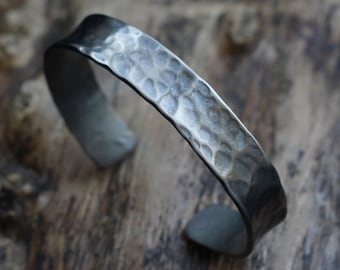 Puede incluir: Un brazalete de metal martillado plateado. El brazalete está hecho de un metal grueso y texturizado y tiene un diseño simple y moderno.
