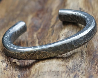 Puede incluir: Un brazalete de metal martillado plateado con un diseño redondeado y abierto. El brazalete está apoyado en una pieza de madera.