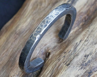 Puede incluir: Un brazalete de metal plateado con un acabado martillado. El brazalete está abierto y tiene una forma redondeada.