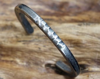 Puede incluir: Un brazalete de plata con acabado martillado. El brazalete está hecho de metal y tiene un diseño simple y moderno.