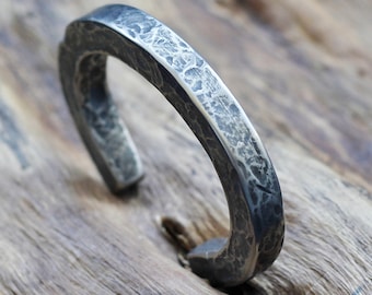 Puede incluir: Un brazalete abierto, de color plateado, con textura martillada. Presenta un aspecto oscuro y envejecido. El brazalete se muestra sobre una superficie de madera, resaltando su estética rústica. Un accesorio único y artesanal.