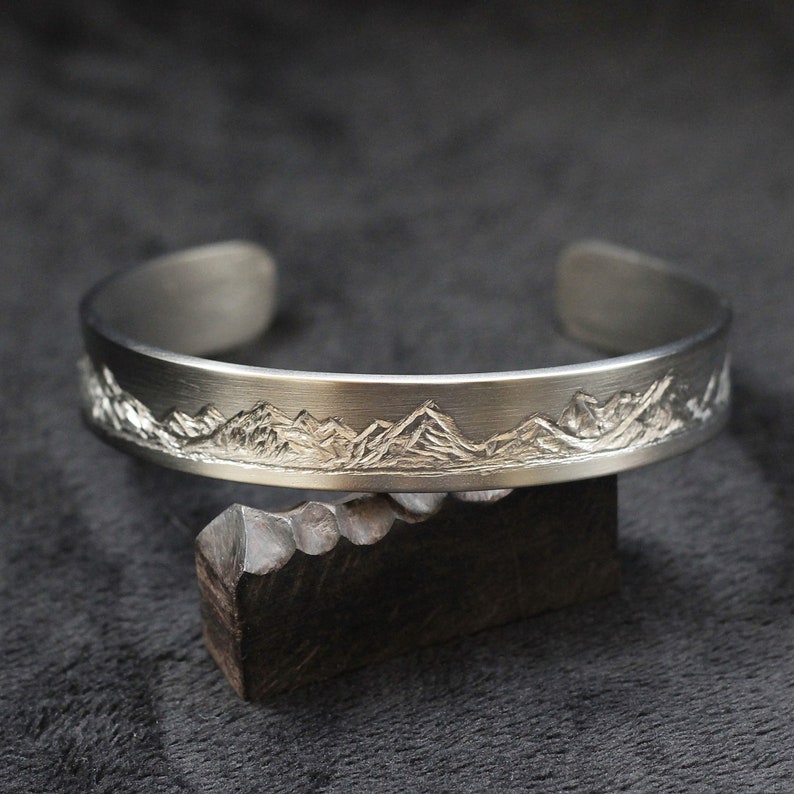 May include: A silver bracelet with a mountain range design engraved on the front.