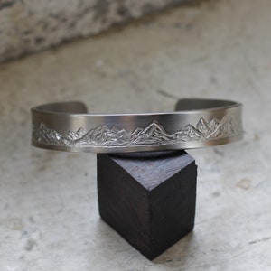 May include: A silver metal cuff bracelet with a mountain range design engraved on the front.