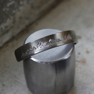 May include: A silver metal band with a mountain range design engraved on the top. The band is sitting on a silver metal surface.