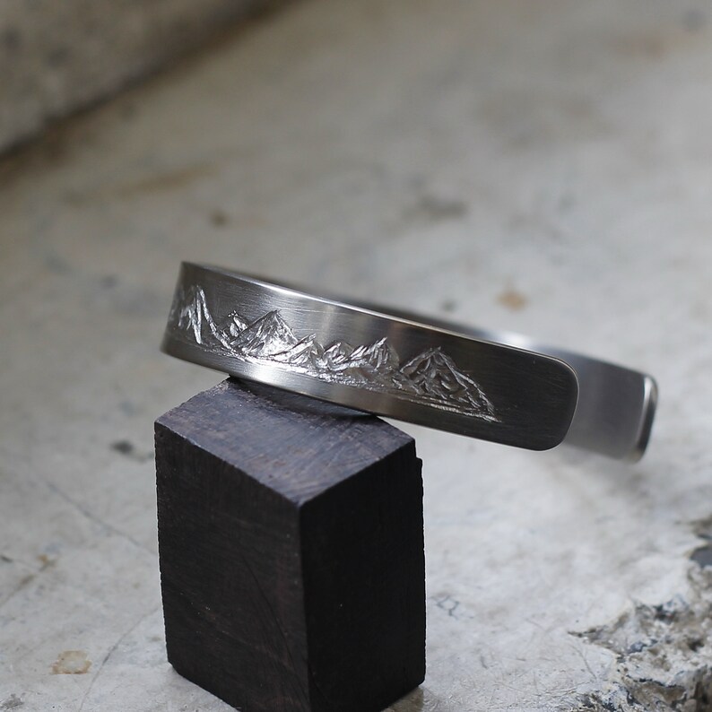May include: A silver bracelet with a mountain range design engraved on the front. The bracelet is on a dark wood block.