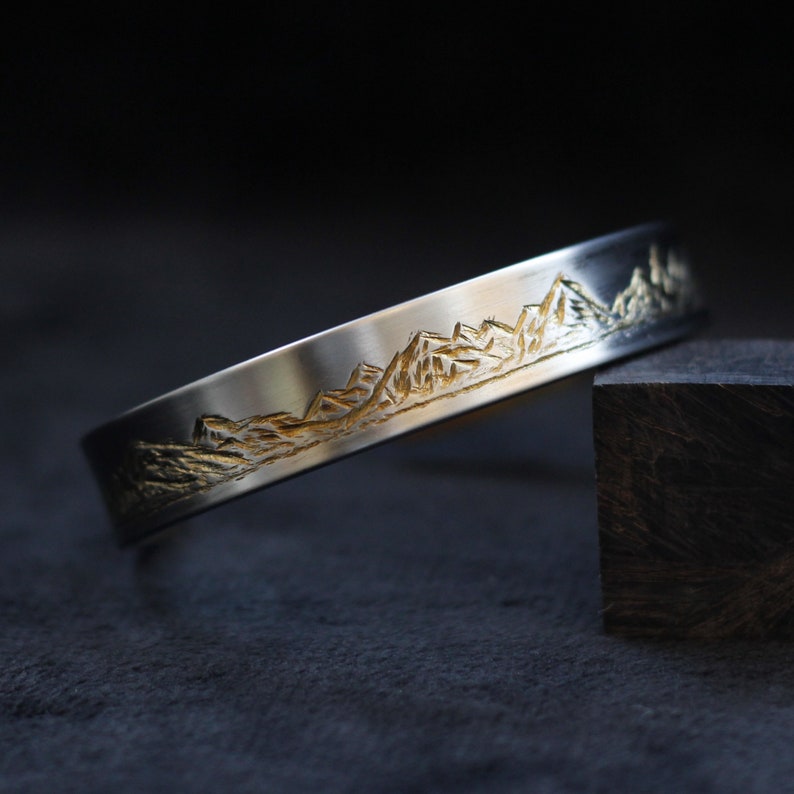 May include: A silver bracelet with a gold mountain range design. The bracelet is on a dark surface.