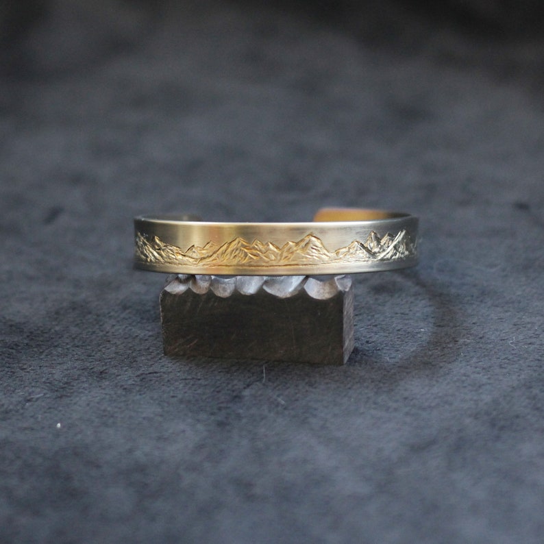 May include: A gold-toned metal bracelet with a mountain range design engraved on the front.