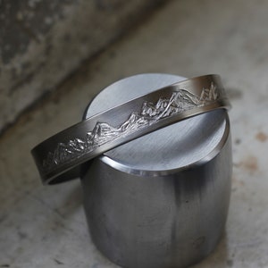 May include: A silver metal bracelet with a mountain range design engraved on the surface.