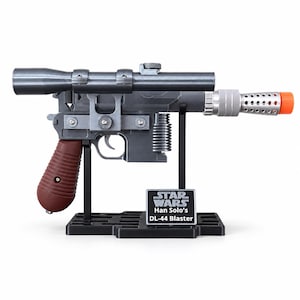 May include: A replica DL-44 blaster with a dark gray finish, a brown ribbed grip, and a long scope mounted on top. The barrel features a silver perforated heat sink and an orange tip. It rests on a black stand with a plaque reading Star Wars Han Solo's DL-44 Blaster.