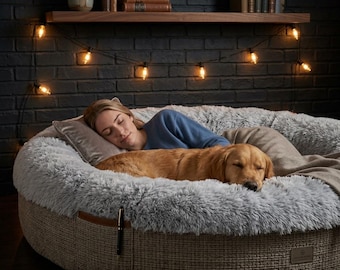 May include: A large, round pet bed with a textured, tan woven exterior and a plush, light gray faux fur interior. The bed features a small brown leather accent tab on the side and a rectangular brand label on the lower front edge.