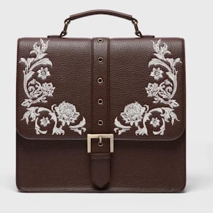 May include: A dark brown structured satchel bag with a pebbled texture, featuring a top handle and a central vertical strap with a metallic buckle. The front flap is decorated with symmetrical white floral embroidery on both the left and right sides.