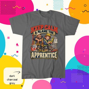 May include: A dark charcoal grey t-shirt featuring a graphic illustration of two construction workers with tools and the text "KEEP CALM & BLAME THE APPRENTICE" in red and white lettering, along with a small sign that reads "NEARLY LEVEL?"
