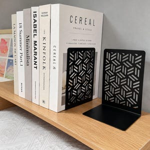 Puede incluir: Dos sujetalibros de metal negro con un patrón geométrico recortado de rectángulos repetidos. Los sujetalibros están colocados sobre un estante de madera clara, sosteniendo una fila de cinco libros de tapa dura con lomos blancos.